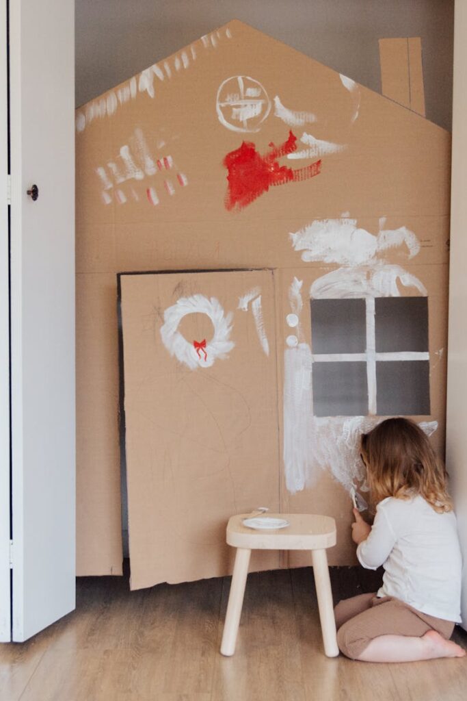 A young girl enjoys painting a cardboard house indoors, emphasizing creativity and fun.